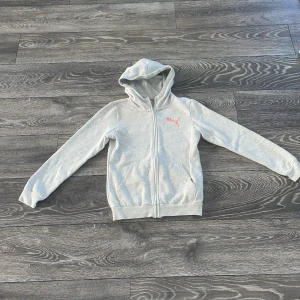 Grå hoodiejacka från Puma - Grå hoodiejacka från Puma med dragkedja framtill och orange Puma-logga på bröstet. Jackan har huva, två fickor och ribbade muddar vid ärmslut och nederkant. Perfekt för chill dagar eller när du vill ha en sportig look.