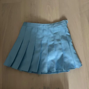 Ljusblå plisserad kortkjol  - Supersöt ljusblå plisserad kortkjol. Det finns två knappar: en unik liten björnformad knapp i midjan och den ursprungliga samt en dragkedja i sidan så storleken kan justeras efter behov (därmed passar den XXS, XS och S). Kjolen har klassisk skoluniform-vibe och är perfekt för dig som gillar en söt och stilren look. 