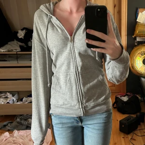 Grå hoodie med dragkedja från H&M Divided - Mysig grå hoodie från H&M Divided i storlek S. Tröjan har huva med snörning, hel dragkedja framtill och mjukt teddyfoder på insidan. Perfekt för kyliga dagar och enkel att matcha med jeans eller leggings.