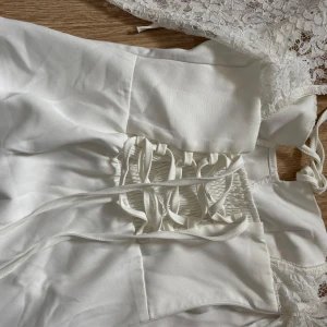 Vit klänning med spetsdetaljer - Elegant vit klänning med långa spetsärmar och snörning i ryggen. Klänningen har en vacker spetsdetalj vid halsen och en figurnära passform. Perfekt för speciella tillfällen.