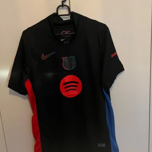 Svart fotbollströja från Nike - Snygg svart fotbollströja från Nike med röda och blå detaljer. Tröjan har korta ärmar och ett tryck på framsidan samt spelarens namn och nummer på baksidan. Perfekt för fotbollsfans som vill visa sitt stöd.