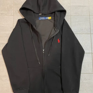 Svart hoodie från Ralph Lauren - Säljer en stilren svart hoodie från Ralph Lauren med dragkedja och huva. Den har en broderad röd logotyp på bröstet och justerbara snören vid huvan. Perfekt för en avslappnad stil. Det står M men det passar S.