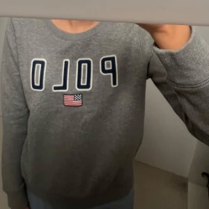Grå sweatshirt från Ralph Lauren - Säljer en grå sweatshirt från Ralph Lauren med 'POLO' och en amerikansk flagga på framsidan. Tröjan har långa ärmar och en rund halsringning. 