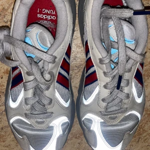 Adidas Yung-1 sneakers storlek 37,5 - Säljer ett par Adidas Yung-1 sneakers i grått med röda och blå detaljer. Skorna har en retro design med snörning och en bekväm passform. Perfekta för en sportig look. 