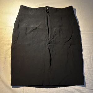 Svart kjol från Karl Lagerfeld - Elegant svart kjol från Karl Lagerfeld med en klassisk design. Kjolen har en diskret dragkedja och knappar framtill för en stilren look. Perfekt för en sofistikerad stil.