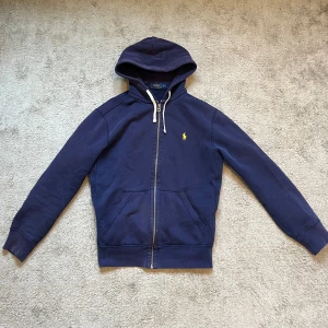 Blå hoodie från Ralph Lauren - Säljer en klassisk blå hoodie från Ralph Lauren med dragkedja. Hyfsat skick förutom att det är ett hål på armbågen därav priset. Priset är fast!