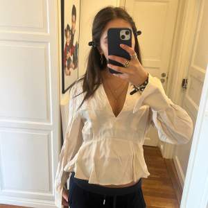 Säljer en stilren vit blus med v-ringning och långa ärmar. Blusen har en lätt volangdetalj vid nederkanten som ger en feminin touch. Perfekt för en chic och avslappnad look.