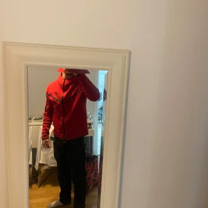 Nike tech röd - Snygg Nike tech i röd med dragkedja och svarta detaljer. Techen har en hög krage och en ficka med dragkedja på ärmen. Perfekt för en sportig look.