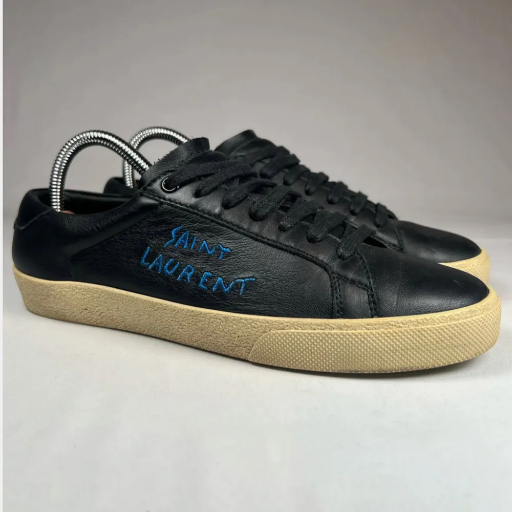 Snygga svarta sneakers från Saint Laurent med blå logotyp på sidan. Skorna har en rund tå och snörning framtill. De har en beige sula som ger en cool kontrast till det svarta skinnet.. Kengät.