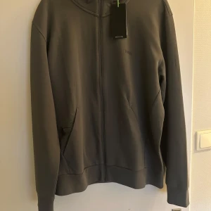 Grön hoodie från BOSS - Snygg och stilren grå hoodie från BOSS med dragkedja framtill. Perfekt för en avslappnad look. Den har långa ärmar och praktiska fickor på sidorna. Ett måste för garderoben!