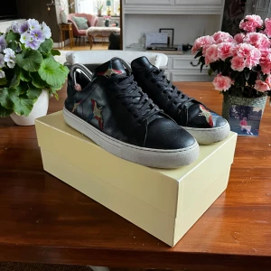 Svarta sneakers från Axel Arigato - Snygga svarta sneakers i skinn från Axel Arigato med färgglada broderade fåglar på sidorna. Skorna har en klassisk snörning och en vit sula som ger en stilren kontrast. Perfekta för att ge din outfit en unik touch.