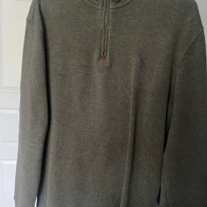 Olivgrön tröja från Ralph Lauren - Säljer en stilren olivgrön tröja från Ralph Lauren med dragkedja och broderad logga i lila på bröstet. Tröjan har långa ärmar och en hög krage, perfekt för kyligare dagar. Passar både till vardags och mer uppklädda tillfällen. Hör av er vid fler frågor!