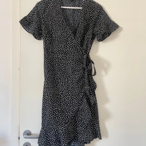 Svart kortärmad klänning med vita prickar - Säljer en söt svart klänning med vita prickar och volangdetaljer. Klänningen har korta ärmar och en omlott-design med knytning i sidan. Perfekt för en somrig look!