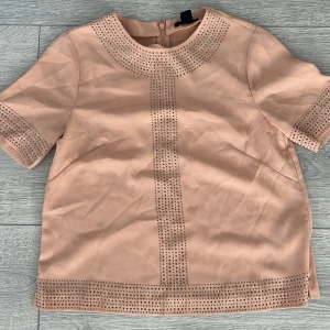 Beige kortärmad topp från H&M - Säljer en elegant beige topp från H&M med korta ärmar och glittriga detaljer. Toppen har en dragkedja i ryggen och är perfekt för en stilren look. Passar bra till både jeans och kjol.