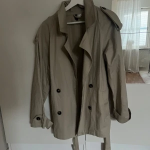 Beige trenchcoat - Snygg beige trenchcoat med knappar och bälte i midjan. Perfekt för vår och höst. Klassisk design med långa ärmar och axelklaffar.