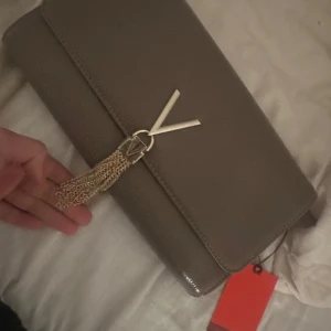 Taupe kuvertväska från Mario Valentino - Elegant taupe kuvertväska från Mario Valentino med en stilren design. Väskan har en dekorativ tofsdetalj och en diskret logotyp på framsidan. Perfekt för att lyfta din outfit med en touch av lyx.