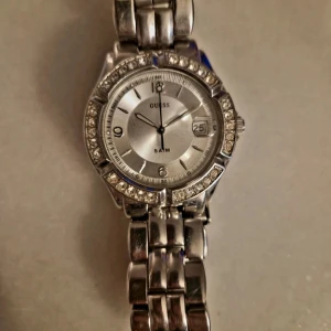 Silverfärgad klocka från Guess - Snygg silverfärgad klocka från Guess med glittrande stenar runt urtavlan. Modell: 75499M1. Vattentät och behöver nytt batteri