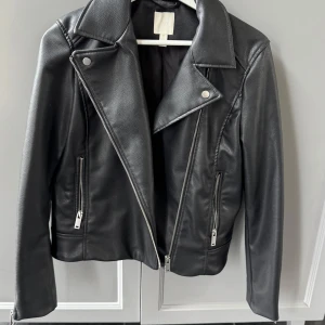 Svart skinnjacka från H&M - Snygg svart skinnjacka från H&M med dragkedja och klassisk krage. Jackan har långa ärmar och två fickor med dragkedja framtill. 