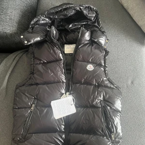 Svart dunväst från Moncler - Snygg svart dunväst från Moncler med dragkedja och avtagbar huva. Västen har en glansig finish och Moncler-logga på bröstet. Qr koden funkar som den ska, nfc funkar också. Den passar både M/L.