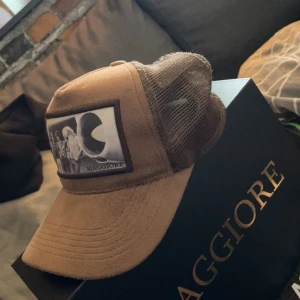 Brun keps från Maggiore - Snygg brun keps från Maggiore med ett svartvitt tryck framtill. Kepsen har ett nätmaterial på sidorna och baksidan för extra ventilation. Perfekt för en avslappnad stil. Den är i Nyskick och köparen får med låda och dustbag 