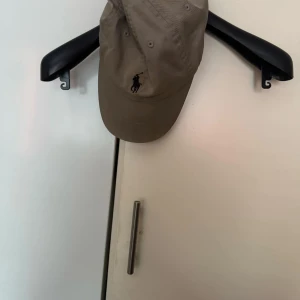 Ralph Lauren - Snygg beige keps från Ralph Lauren med den klassiska loggan broderad i svart framtill. Kepsen är tillverkad i 100% bomull och har en justerbar rem baktill för perfekt passform.