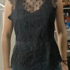 Svart spetsblus - Elegant svart blus med spetsdetaljer och transparenta partier. Blusen har en vacker spetskrage och är ärmlös, vilket ger en sofistikerad look. Har aldrig använt den. Vet inte riktigt vilken storlek den är, men den är ganska stor för mig så jag kunde säga att den är typ storlek 38/M. 