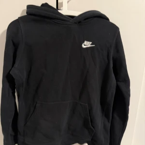 Svart hoodie från Nike - Säljer en klassisk svart hoodie från Nike med vit logga på bröstet. Tröjan har en stor ficka framtill och en bekväm huva. Perfekt för en avslappnad stil. Passar både till vardags och träning.