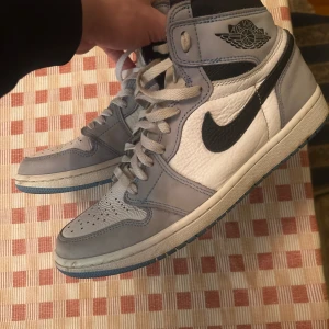 Nike Air Jordans 1 unc blue - Snygga Nike Air Jordans i en stilren kombination av grått och vitt med svarta detaljer. Skorna har en klassisk snörning och rund tå. Perfekta för dig som älskar sneakers med en ikonisk design.