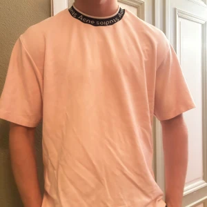 Rosa t-shirt från Acne Studios - Säljer en stilren rosa t-shirt från Acne Studios med korta ärmar och en svart rundad krage med vit text. Perfekt för en avslappnad look.