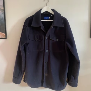 Mörkblå overshirt i polyester - Köpte för några år sen, använde den ett fåtal gångar så skicket är 9.5/10. Vill endast bli av med den då den tar plats och inte används . Stilren mörkblå overshirt från Jack & Jones i 100% polyester. Jackan har långa ärmar och knappar framtill samt två bröstfickor med knappar. Perfekt för lager-på-lager-stil.