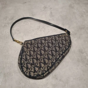 Vintage Dior mini saddle bag / pochette - En vintage Dior Mini Saddle Bag/Pochette säljes. Blå och beige/brun (ser något mer grå ut på bilderna men är i det klassiska monogram-tyget). Tunt handtag med gulddetaljer. Vintage och använd men i mycket fint skick!  Fler bilder kan skickas om så önskas. Skickas mot frakt eller hämtas i Örebro.