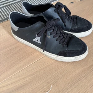 Svarta sneakers från Axel Arigato - Snygga svarta sneakers från Axel Arigato med vit sula och grå detaljer. Skorna har snörning och en dekorativ bokstav 'A' på sidan. Perfekta för en stilren look.