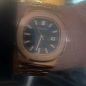 Guldfärgad klocka med svart urtavla - Elegant guldfärgad klocka med svart urtavla och datumvisning. Klockan har en robust länkarmband och tydliga timmarkeringar. Perfekt för den som vill ha en stilren accessoar.