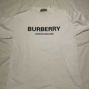 Vit t-shirt från Burberry - Säljer en stilren vit t-shirt från Burberry med svart text 'BURBERRY LONDON ENGLAND' på framsidan. Perfekt för en casual look med en touch av lyx. Passar bra till jeans eller shorts.