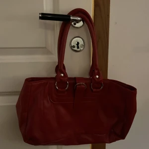 Röd axelväska i syntet - Säljer en snygg röd axelväska med dubbla handtag och silverfärgade metalldetaljer. Väskan har en klassisk rektangulär form och är tillverkad i syntetmaterial. Perfekt för dig som vill ha en färgklick till din outfit!