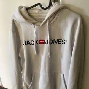 Vit hoodie från Jack & Jones - Säljer en vit hoodie från Jack & Jones med svart logga och röd detalj på bröstet. Tröjan har huva med dragsko och en stor magficka. Perfekt för en avslappnad stil. 