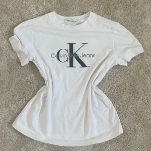 Vit t-shirt från Calvin Klein Jeans - Vit t-shirt från Calvin Klein Jeans med stor CK-logga framtill. Klassisk rund halsringning och korta ärmar. Perfekt basic med stilren design och mjukt bomullsmaterial.