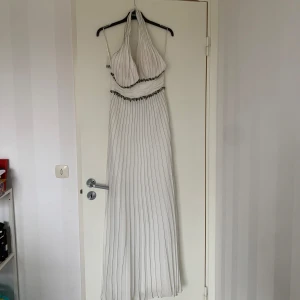 Vit långklänning med halterneck och pärldetaljer - Säljer en elegant vit långklänning med halterneck och fina pärldetaljer vid midjan och bysten. Klänningen har plisserad kjol och öppen rygg, perfekt för speciella tillfällen. 