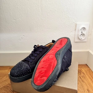 Christian Louboutin navy - Säljer ett par svarta sneakers från Christian Louboutin med svarta nitar på tån och röd sula. Skorna har snörning och är gjorda i mocka med svarta detaljer. Perfekta för dig som vill sticka ut med en lyxig touch.