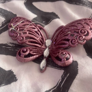 Glittrig fjärilsaccessoar i rosa - Säljer en dekorativ accessoar i form av en fjäril i glittrig rosa färg. Fjärilen har utskurna detaljerade vingar och tre stora, facetterade stenar på kroppen. Hängs upp med en tunn silverfärgad tråd.