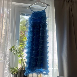 Blå volangklänning från YAS - Säljer en blå axelbandsklänning från YAS med volanger över hela klänningen. Klänningen är vadlång och har en luftig passform med tunna axelband. Perfekt för sommarens tillfällen när du vill sticka ut med en unik look. Dock ser man att den blivit lite defekt på ryggen efter en sittning, vet inte varför den är sliten just det men syns på bild 5. 