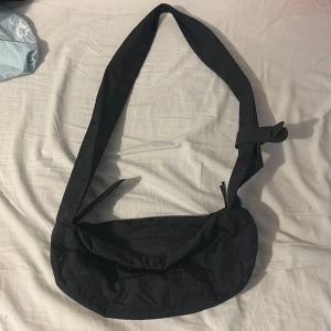 Svart axelväska från Weekday - Säljer en svart axelväska från Weekday med justerbart band och dragkedja upptill. Väskan har en enkel, stilren design och är tillverkad i ett lätt syntetmaterial. Perfekt för att bära med sig det viktigaste i vardagen.