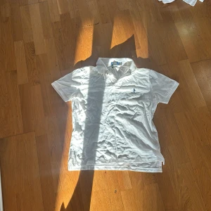 Vit piké från Polo Ralph Lauren - Klassisk vit pikétröja från Polo Ralph Lauren med broderad logga på bröstet. Tröjan har korta ärmar, krage och knappar vid halsen. Perfekt för en stilren och sportig look.