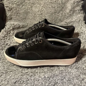 Svarta sneakers från Lanvin med lackad tå - Säljer ett par svarta sneakers från Lanvin med snygg lackad tå och vit platt sula. Skorna har snörning och är tillverkade i mocka med detaljer i skinn. Perfekta för dig som gillar stilrena och exklusiva sneakers.