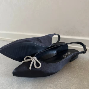 Svarta ballerinaskor med rosett från Miss F - Snygga svarta ballerinaskor från Miss F med spetsig tå och dekorativ glittrig rosett framtill. Skorna har öppen häl med rem och låg platt sula. Perfekta för dig som vill ha en elegant och stilren look.