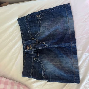 Lågmidjad mini kort jeanskjol med fickor - Säljer en klassisk blå mini jeanskjol med två framfickor och två bakfickor.  Kjolen har dragkedja och dubbla knappar framtill samt kontrastsömmar. Perfekt för dig som gillar en tidlös denimlook. Passar S och XS