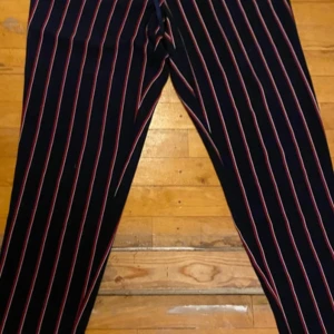 Svarta kostymbyxor med röda och vita ränder från Tommy Hilfiger - Säljer ett par svarta kostymbyxor från Tommy Hilfiger med tunna röda och vita vertikala ränder. Byxorna har klassisk passform och är perfekta för en stilren look. Snygga att matcha med en enkel topp eller skjorta.