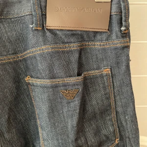 Mörkblå jeans från Emporio Armani - Snygga mörkblå jeans från Emporio Armani med klassisk femficksdesign och logotyp på bakfickan. Jeansen har orange kontrastsömmar och läderpatch med märke i midjan. Perfekta för dig som gillar stilrena och exklusiva jeans.