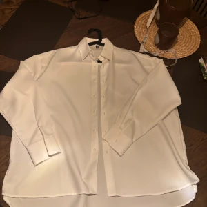 Vit skjorta från ASOS - Klassisk vit skjorta från ASOS med lång ärm och knappar framtill. Skjortan har en rak passform och är tillverkad i ett slätt, mjukt material som ger en stilren look. Perfekt att styla till olika outfits. Storlek oversized liknande L