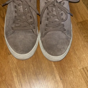 Bruna sneakers i mocka - Säljer ett par bruna sneakers i mocka med snörning och rund tå. Skorna har en vit platt sula och brunt innerfoder. Perfekta för dig som gillar en stilren och enkel look. Storlek 45 men passar 44. 